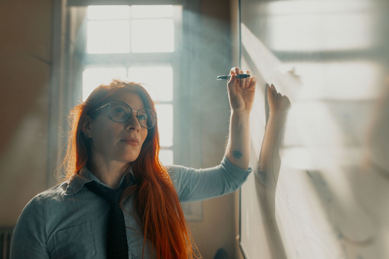 Young woman writing on a whiteboard in a sunlit classroom, focused and reflective.