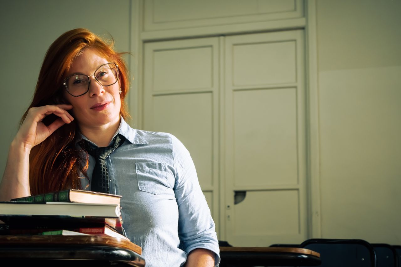 A confident woman with glasses sits in a classroom, surrounded by books, exuding intelligence and curiosity.